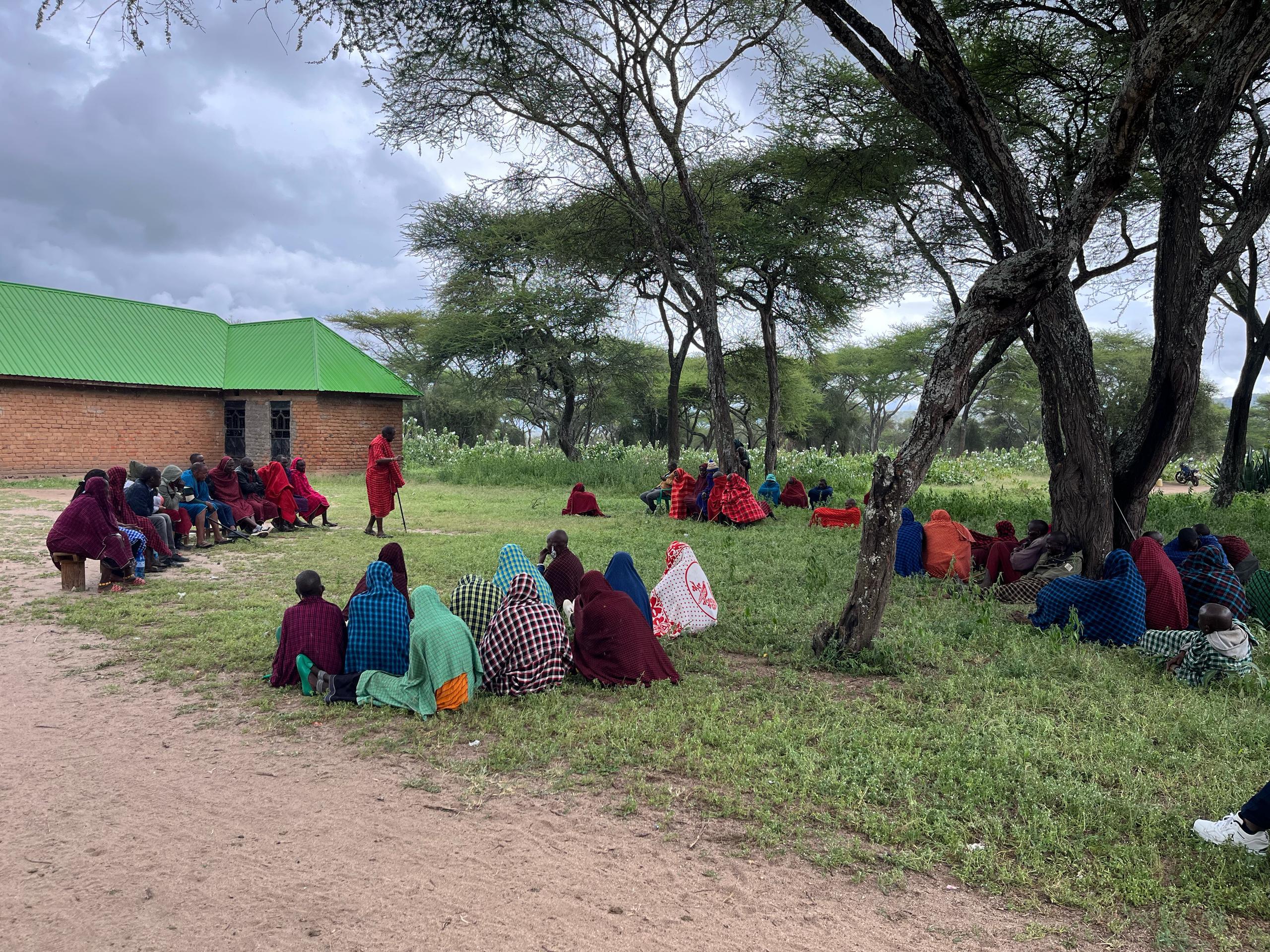 A group of people in seated under large trees, as the community meeting goes on.